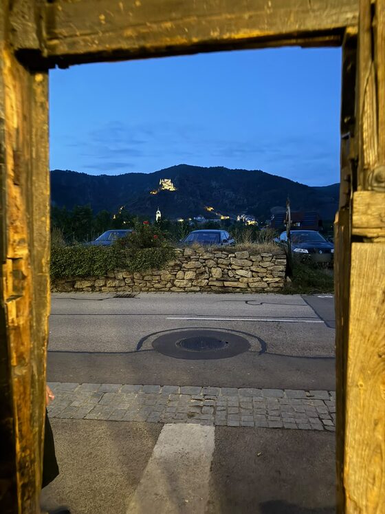 Blick durch den hölzernen Türrahmen zur blauen Stunde über Straße und Steinmauer auf die angestrahlte Ruine Dürnstein